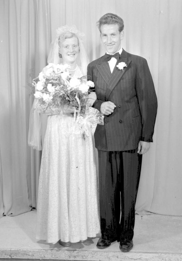 Ein Mann und eine Frau stehen nebeneinander auf einer Bühne und lächeln, wobei die Frau einen Blumenstrauß hält und ein Vorhang hinter ihnen zu sehen ist.