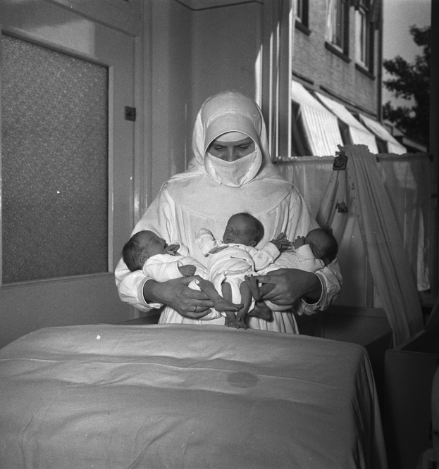 Eine Frau in einem Krankenhausbett hält zwei Babys im Arm, umgeben von einer Wand, einer Tür, einem Fenster, einem Baum und einem Gebäude im Hintergrund, dargestellt in Schwarz-Weiß.