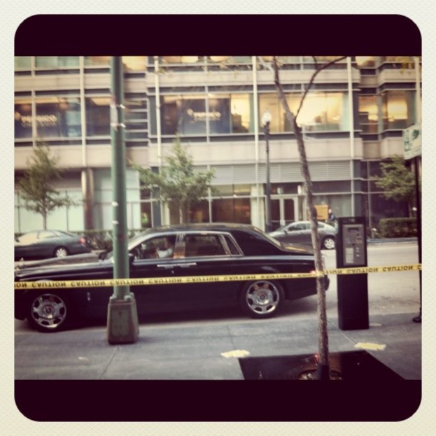 Ein Auto ist auf dem Bild in der Mitte mit Pfählen und Bäumen, einem Gebäude, Fahrzeugen und Pflanzen im Hintergrund und einem Weg am Boden geparkt.