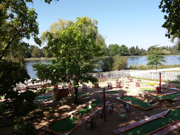 Minigolfanlage in einem Park mit grünem Gras, Bäumen und einem Zaun, umgeben von verstreuten Stühlen und Tischen, mit einem Wasserlauf und einem Boot im Hintergrund unter einem sichtbaren Himmel.