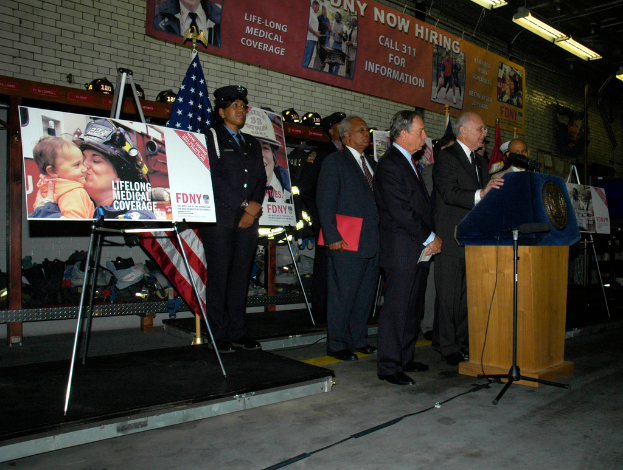 Eine Gruppe von Menschen in der Nähe eines Podiums mit Mikrofonen, Schildern auf Ständern, Flaggen und Schildern an einer Wand, alles unter hellen Lichtern.