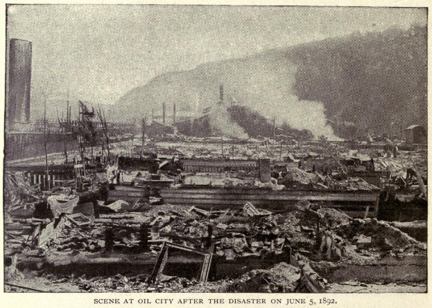 Schwarzes und weißes Foto einer Stadt nach einer Katastrophe am 5. Juni 1892, das Gebäude, Mäste, Rauch und einen Hügel im Hintergrund zeigt, mit Text unten.