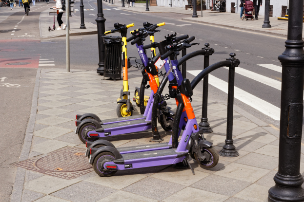 Eine Reihe von Elektro-Scootern, die entlang einer Straße geparkt sind, mit Fußgängern, Radfahrern, einem Müllcontainer im Vordergrund und Gebäuden, Bäumen und Pflanzen im Hintergrund.