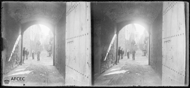 Ein Schwarz-Weiß-Foto einer Gruppe von Menschen, die durch einen Tunnel mit Wänden auf beiden Seiten und Gebäuden im Hintergrund gehen, mit einem Wasserzeichen, das darauf hinweist, dass es eine stereoskopische Ansicht der Stadt Catania, Sizilien, ist.