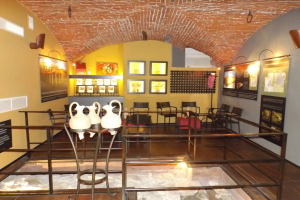 Ein Museumsraum voller Tische, Stühle, eines Geländers, Vasen, Texttafeln, Wandrahmen, Lampen und diverser anderer Ausstellungsstücke.