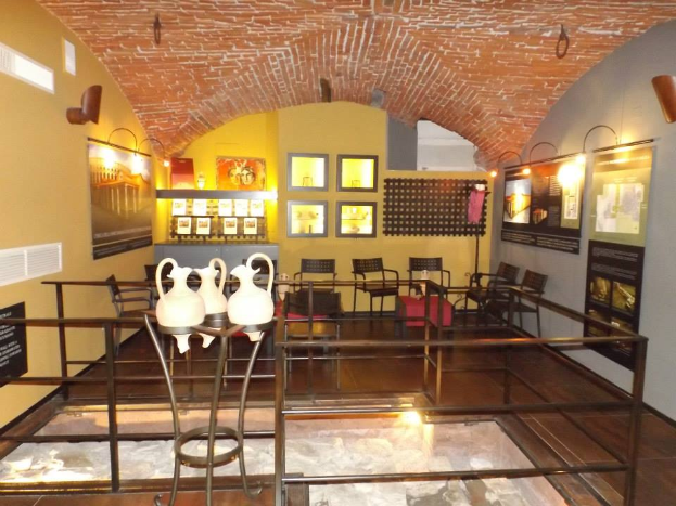 Ein Museumsraum voller Tische, Stühle, eines Geländers, Vasen, Texttafeln, Wandrahmen, Lampen und diverser anderer Ausstellungsstücke.