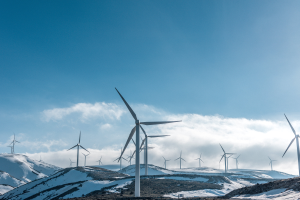 Eine Gruppe von Windkraftanlagen in einem verschneiten Feld mit schneebedeckten Hügeln und Wolken im Hintergrund, wahrscheinlich Teil eines Erneuerbare-Energie-Projekts.