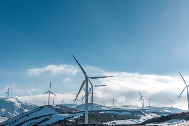 Eine Gruppe von Windkraftanlagen in einem verschneiten Feld mit schneebedeckten Hügeln und Wolken im Hintergrund, wahrscheinlich Teil eines Erneuerbare-Energie-Projekts.