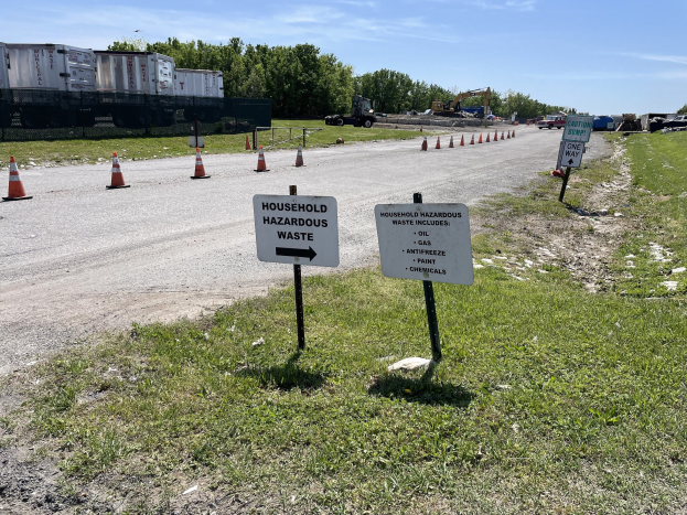 Ein Straßenschild für 'Haushaltsgefährliche Abfälle' umgeben von Gras, Verkehrskegeln, Fahrzeugen, Bäumen und einem bewölkten Himmel.