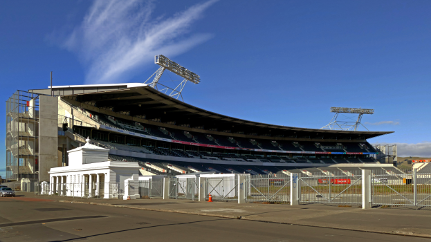 Ein großes Stadion mit zahlreichen parkenden Autos davor, umgeben von einer Straße, einem Zaun, Pfosten, Laternen und Schildern, mit Bäumen und einem bewölkten Himmel im Hintergrund.