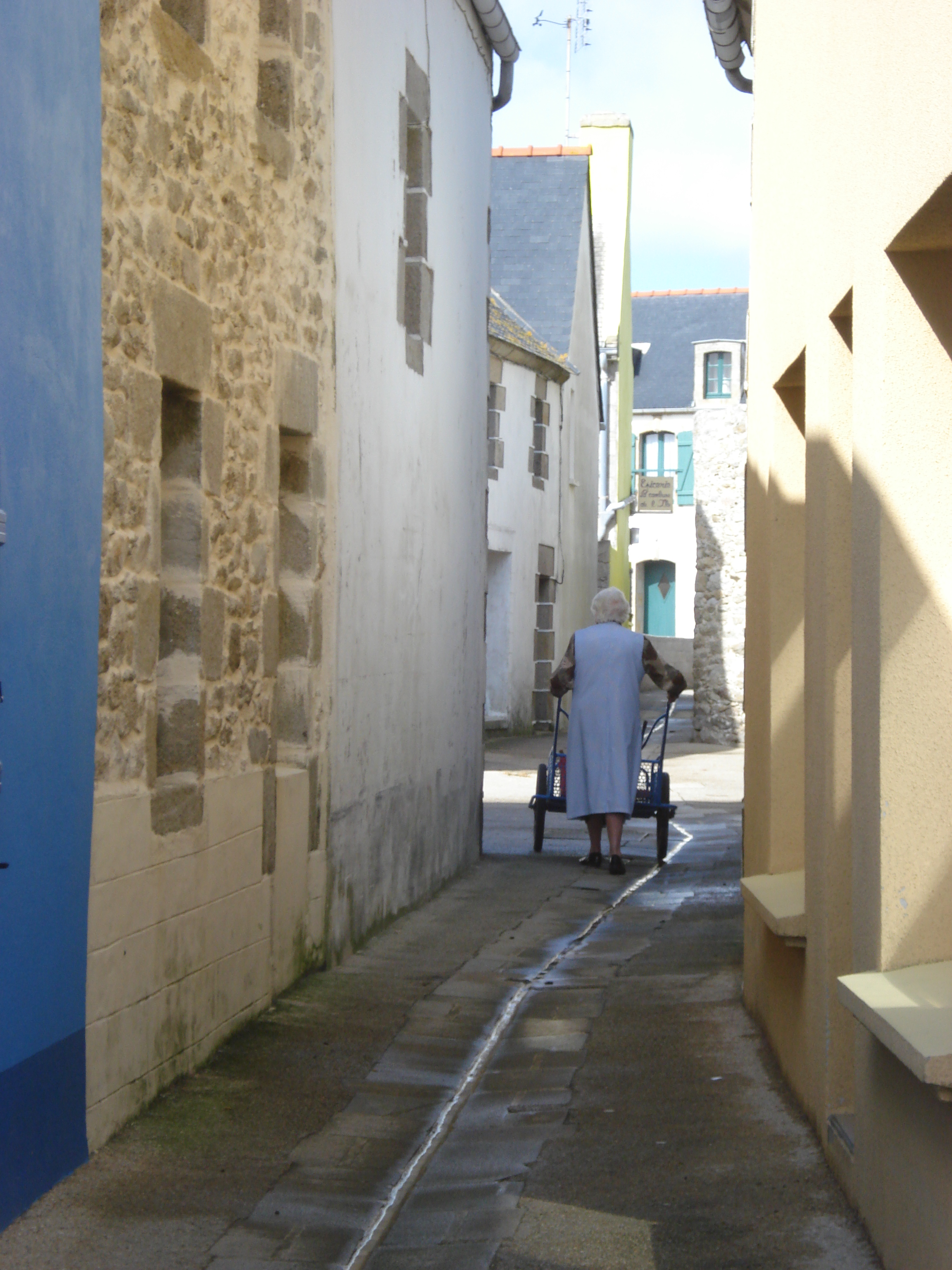 Eine ältere Frau geht mit einem Einkaufswagen durch eine enge Gasse, flankiert von Gebäuden auf beiden Seiten, mit weiteren Gebäuden und dem Himmel im Hintergrund.