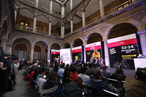 Eine Gruppe von Menschen sitzt auf einem Pressetermin für das CDMX 2017-Event, mit einem Rednerpult und Mikrofon in der Mitte, umgeben von einer Menge mit Kameras, Schildern, Säulen, Geländern, Fenstern und einer Wand im Hintergrund.