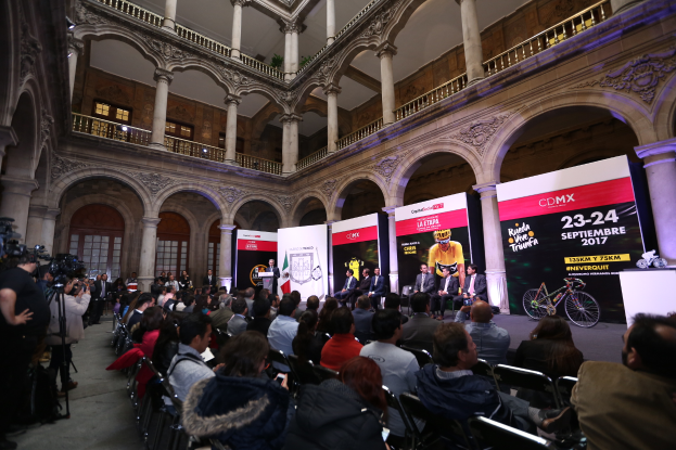 Eine Gruppe von Menschen sitzt auf einem Pressetermin für das CDMX 2017-Event, mit einem Rednerpult und Mikrofon in der Mitte, umgeben von einer Menge mit Kameras, Schildern, Säulen, Geländern, Fenstern und einer Wand im Hintergrund.