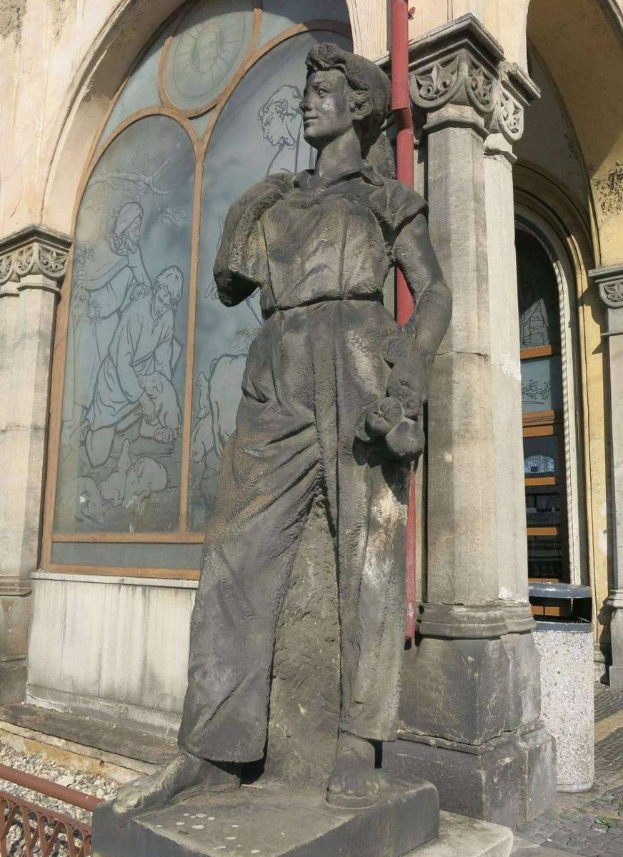 Eine Statue einer Frau steht vor einem kirchenähnlichen Gebäude mit einem Glasfenster, Säulen und einem roten Pfahl, das likely als Holocaust-Mahnmal dient.