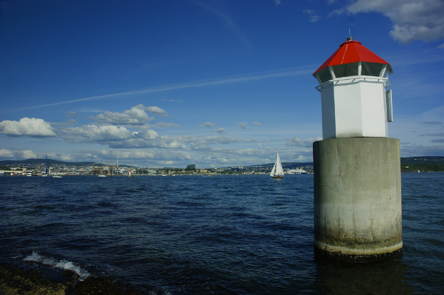 Boote auf dem Meer mit einem Leuchtturm im Vordergrund, Gebäude, Berge und den Himmel im Hintergrund.