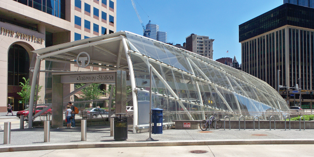 Eine belebte Stadtstraße mit hohen Gebäuden, einem Bushaltestellen, Fußgängern, Fahrzeugen, einem Metallzaun, einem Mülleimer, Strommasten, einem Schild, einer Ampel, Bäumen und einem bewölkten Himmel.