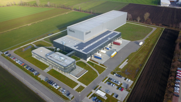 Luftaufnahme eines großen Industriegebäudes, wahrscheinlich eine Fabrik, auf einem Feld umgeben von Bäumen, Fahrzeugen, Straßen und Masten, mit saftigem grünem Gras.