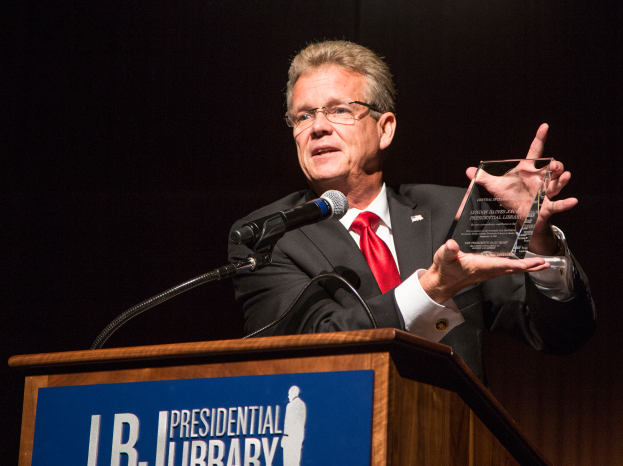 Mann in Anzug und Krawatte steht an einem Rednerpult und hält eine Trophäe in der Hand, während er eine Auszeichnung während einer Bibliothekspreisverleihung erhält.