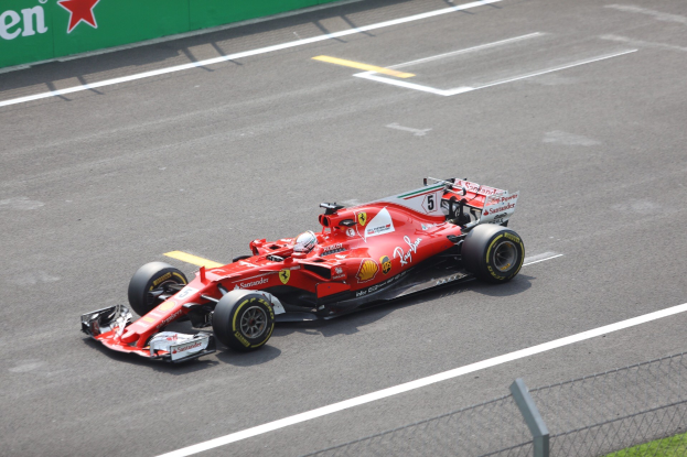 Roter Ferrari Formel 1 Rennwagen, gefahren von Fernando Alonso auf einer Rennstrecke, umgeben von einem Zaun und Gras, mit einem sternmarkierten Sponsorboard im Hintergrund.