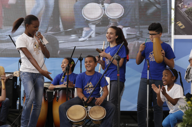 Kinder, die auf der Bühne mit Musikinstrumenten spielen, eine Frau mit einem Mikrofon, Mikrofonständer, eine Banner im Hintergrund und Topfpflanzen rechts.