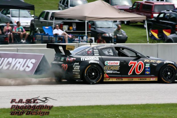 Ein schwarzer Rennwagen auf einer Straße mit Gras am Boden, ein Schild mit Text links daneben, Menschen unter Zelten sitzend und andere Autos im Hintergrund.