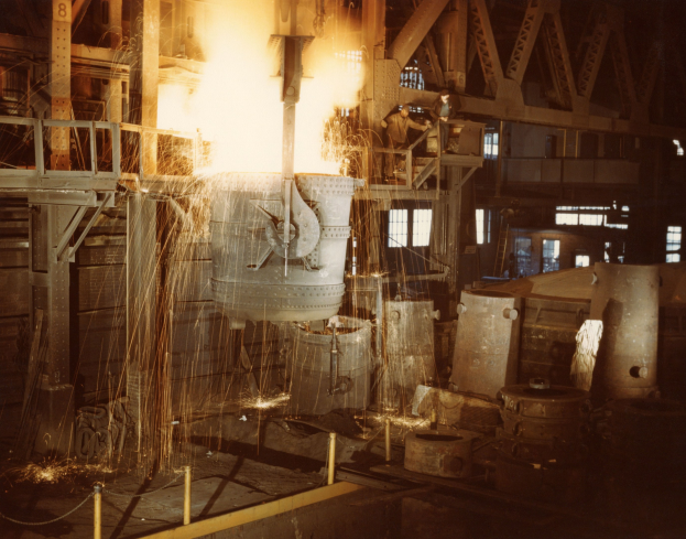 Großes Metallobjekt wird in einen Ofen gegossen, Menschen stehen drumherum, Feuer erhellt die Gegend, Metallobjekte, Pfosten und Fenster im Hintergrund.