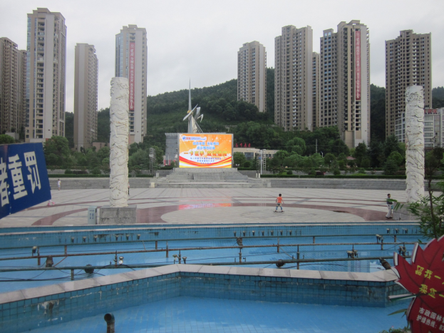 Großer Swimmingpool in städtischer Umgebung mit hohen Gebäuden, wenigen Menschen in der Nähe, einem Banner links, Bäumen und einem klaren blauen Himmel.