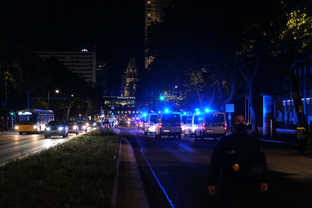 Eine Stadtstraße bei Nacht mit Polizeiwagen, Bussen, Fußgängern auf dem Gehweg, Gras, Bäumen, Gebäuden und Laternen.