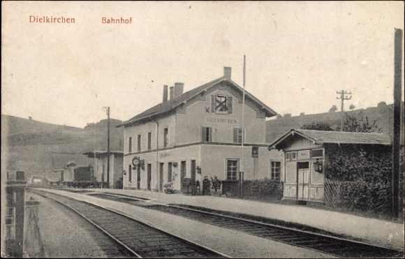 Schwarzes und weißes Foto der Bahnstation Dielkirchen in Deutschland mit einem Zug auf den Gleisen, umgeben von Häusern, Strommasten, Bäumen, Menschen und Bergen sowie einem klaren Himmel im Hintergrund.