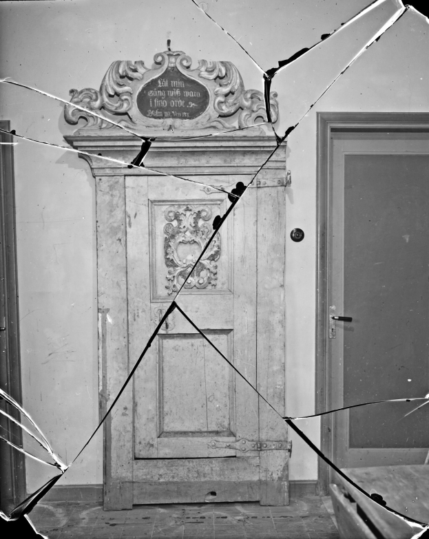 Ein Schwarz-Weiß-Foto einer zerbrochenen Tür in der Mitte eines Raumes mit verstreuten Glasscherben und einer Wand im Hintergrund.