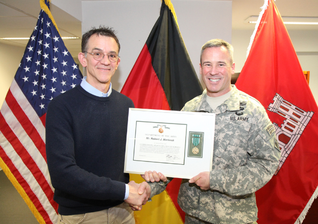 Zwei Männer halten ein Zertifikat mit Flaggen im Hintergrund bei hellem Licht.