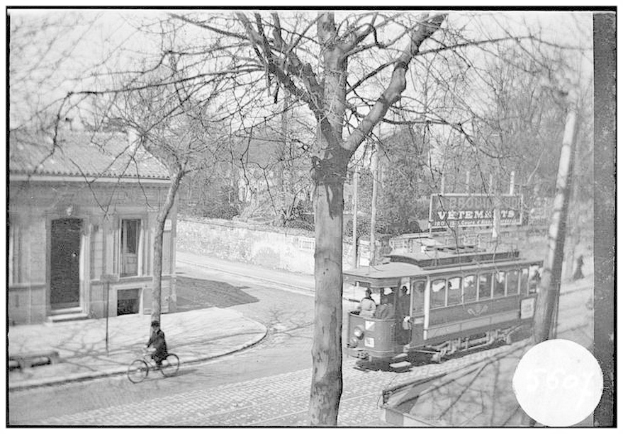 Ein Schwarz-Weiß-Foto einer Straßenbahn auf einer Stadtstraße, mit einem Fahrradfahrer vorne, Passagieren in der Straßenbahn und Bäumen und einem Gebäude im Hintergrund.