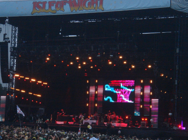 Eine Bühne mit Lichtern, einem Bildschirm und Musikern, die Instrumente spielen, mit einer Fahne oben, einer Menge unten und Fahnen mit Stangen und Ballons.