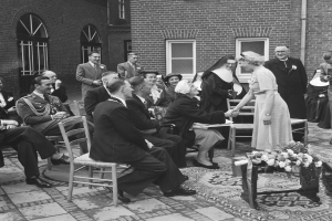 Ein Schwarz-Weiß-Foto einer Hochzeitszeremonie vor einem Gebäude, mit sitzenden und stehenden Gästen und einem Blumenstrauß im Vordergrund, das Gebäude hat Fenster und eine Leitung an der Wand.