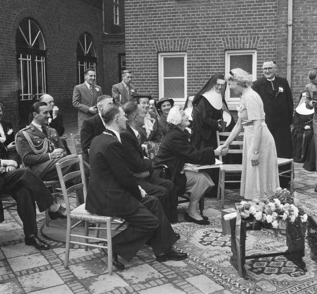 Ein Schwarz-Weiß-Foto einer Hochzeitszeremonie vor einem Gebäude, mit sitzenden und stehenden Gästen und einem Blumenstrauß im Vordergrund, das Gebäude hat Fenster und eine Leitung an der Wand.