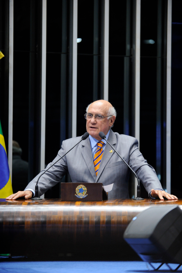 Eine Person, die an einem Rednerpult mit Mikrofonen steht, ein Kasten im Vordergrund und eine Flagge an der linken Wand im Hintergrund.