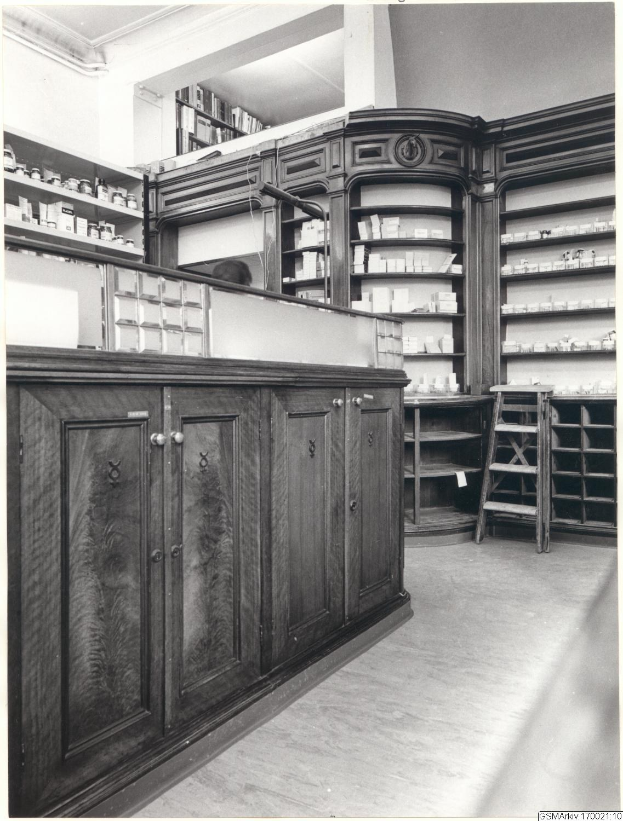 Schwarz-weiß-Foto eines vintage-Apothekeninneren mit einer Theke, Schrankw├Ąnden, einer Leiter, Regalen und Text überall.