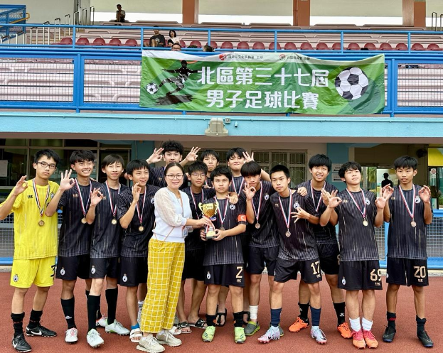 Gruppe junger Männer in Fußballtrikots auf einem Feld, die einen Pokal halten und Medaillen tragen, mit einem Banner "Yokohama U-16 Boys Soccer Team" im Hintergrund und Zuschauern in der Nähe.