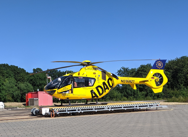 Gelber Hubschrauber auf einem Flughafenvorfeld mit Bäumen und einem klaren blauen Himmel, umgeben von Geräten, die wahrscheinlich für Luftrettungsdienste in Deutschland verwendet werden.