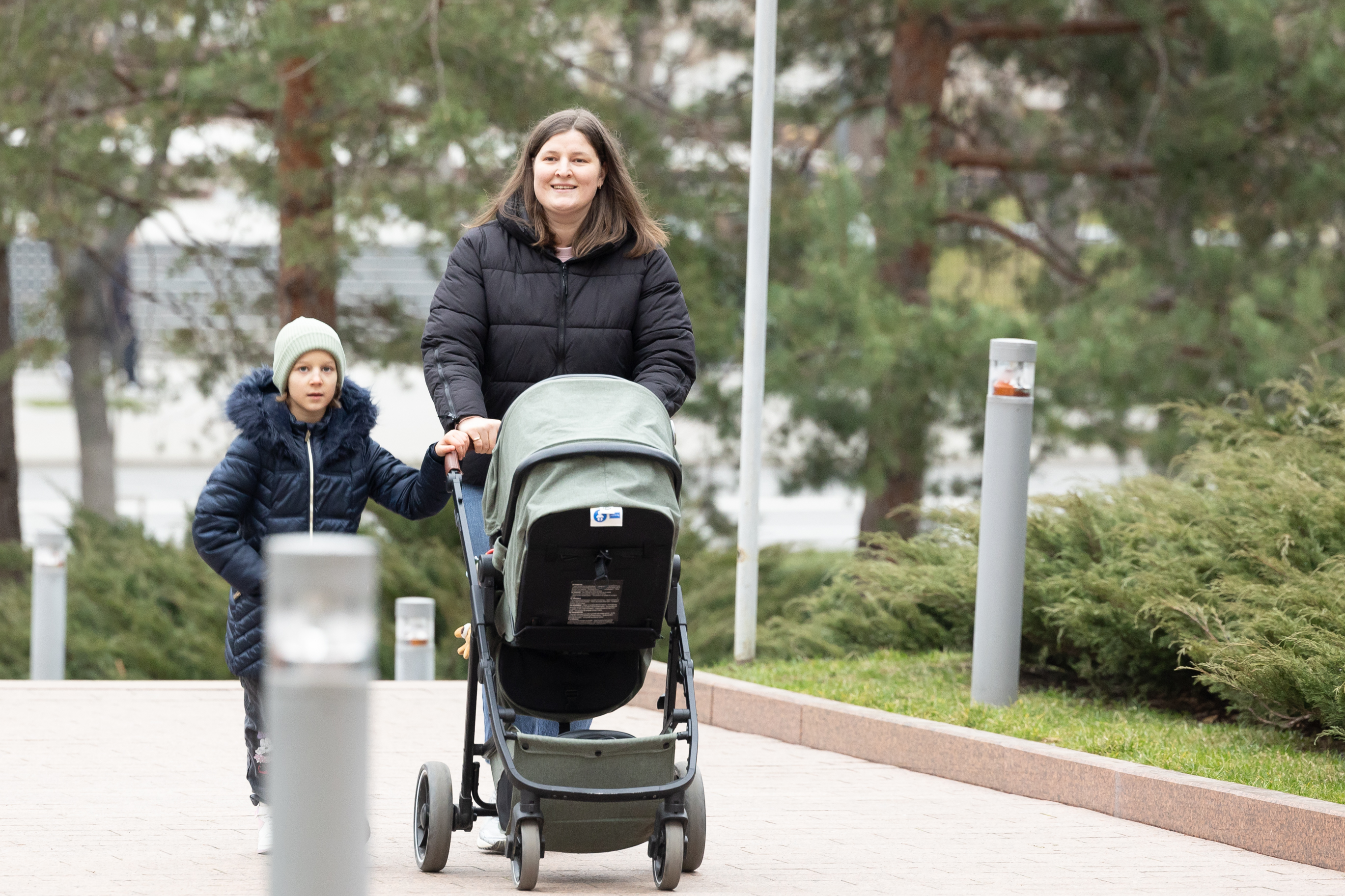 Eine Frau schiebt einen Kinderwagen mit einem Kind eine von Pfählen, Gras, Pflanzen und Bäumen gesäumte Gehweg entlang.