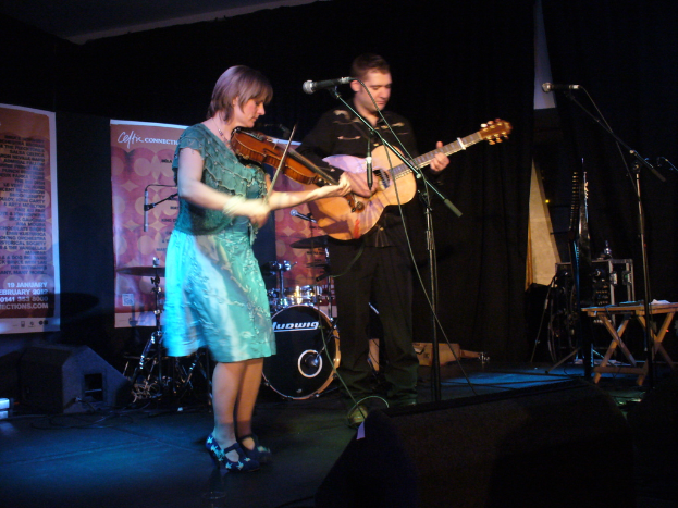 Ein Mann in schwarzem Hemd und Hose spielt Gitarre auf der B├╝hne, begleitet von einer Frau in einem blauen Kleid, die Geige spielt, mit verschiedenen Musikinstrumenten und Lautsprechern im Hintergrund.