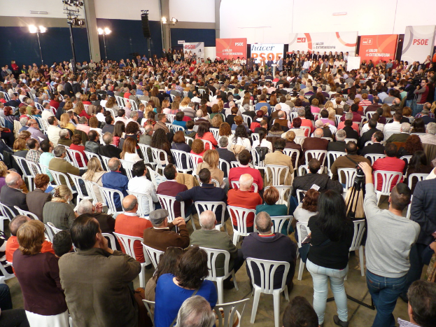 Ein volles Stadion mit vielen Sitz- und Stehplätzen, eine Bühne mit zwei Rednern auf einem Podium und zahlreiche Banner und Lichter.