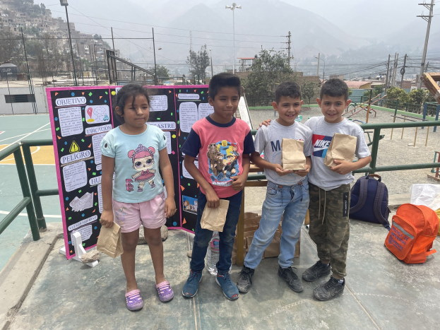 Gruppe von Kindern auf einem Basketballfeld mit Papiertüten in den H├Ąnden, mit Tüten und Gegenst├Ąnden auf der rechten Seite und einer Tafel mit Text auf der linken Seite, vor einem Hintergrund aus Z├Ąunen, Masten, Stra├čenlaternen, B├Ąumen, Geb├Ąuden, H├Ąngen und Himmel.