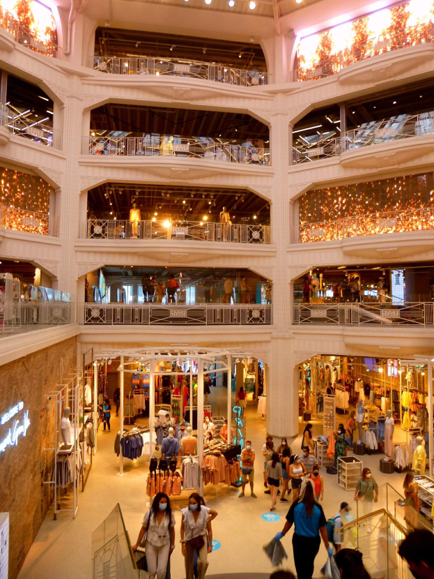 Ein belebter Einkaufszentrumsvorplatz mit Menschen, Schaufensterpuppen mit Kleidung, Geländern, Lampen und Wandtafeln.