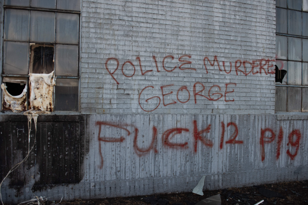 Ein Gebäude mit der Graffiti-Beschriftung "Polizei M"rdert George Fuck 12 P19" an der Seite, das Fenster und verschiedene Gegenstände vorne zeigt.