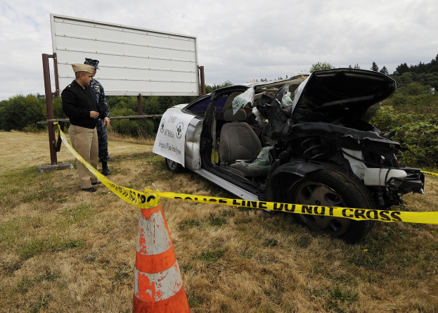 Ein Polizeibeamter in Uniform steht neben einem schwer beschädigten Auto am Straßenrand, mit eingedellter Motorhaube und zersplitterten Fenstern, neben einem Verkehrskegel und einer markierten Stange, unter einem bewölkten Himmel mit Bäumen im Hintergrund.