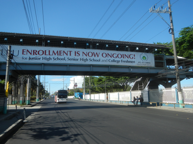 Ein Bus fährt unter einer Brücke mit einem Schild vorbei, auf dem "Jetzt Anmeldung" steht, während Menschen auf einem Fußweg darunter gehen, mit Strommasten und Drähten an der Straße und Bäumen und einem klaren blauen Himmel im Hintergrund.