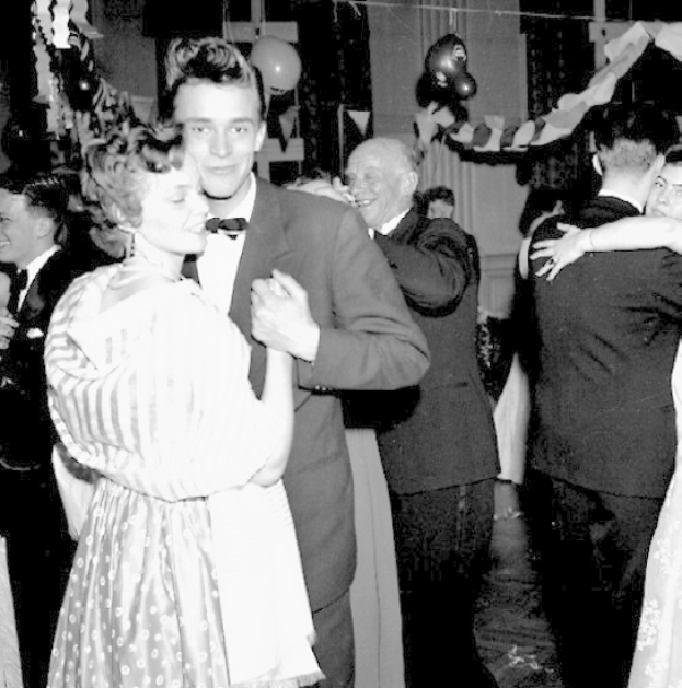 Ein Schwarz-Weiß-Foto von einem Mann und einer Frau, die auf einer Party tanzen, umgeben von lächelnden Menschen, mit Ballons und Deko im Hintergrund.