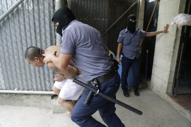 Ein Mann in Gewahrsam wird von zwei Polizeibeamten vor einem Gebäude eskortiert, wobei einer der Beamten seinen Arm hält und der andere einen Schlagstock hält, während eine Wand, eine Tür, ein Rohr und ein Netz im Hintergrund zu sehen sind.