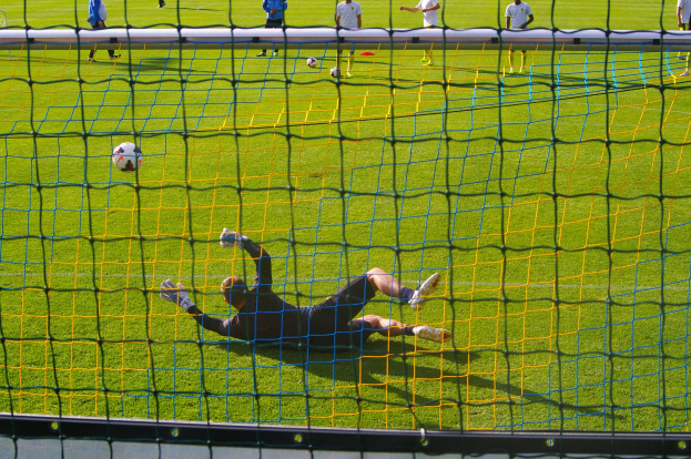 Ein Fußballspieler hält vor einem Tor, mit einem Netz im Vordergrund und einigen Menschen im Hintergrund, sowie mehreren Bällen, die auf dem Boden verstreut sind.